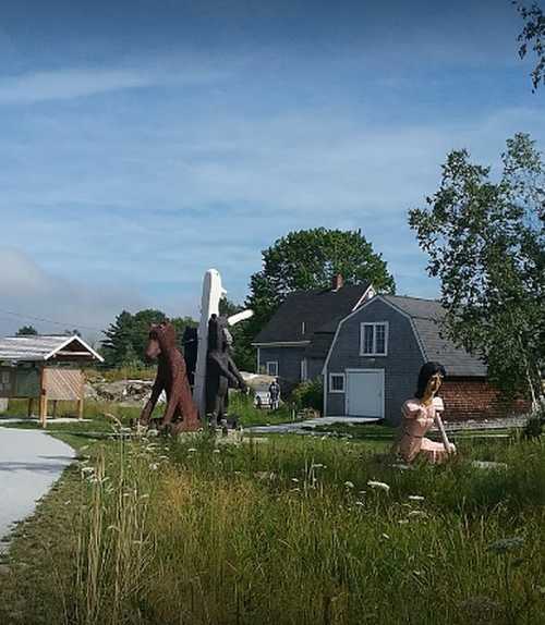 Two large wooden sculptures of a bear and a person in a grassy area near a house and trees under a clear sky.