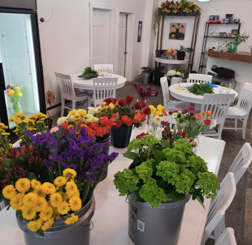 Brightly colored flowers in buckets on tables in a well-lit room with white chairs and shelves.
