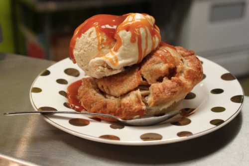 A slice of apple pie topped with a scoop of vanilla ice cream and drizzled with caramel sauce on a polka dot plate.