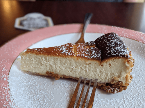A slice of cheesecake on a pink plate, dusted with powdered sugar, with a fork resting beside it.