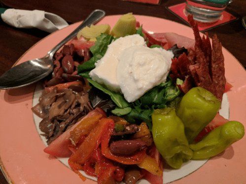 A colorful salad with greens, sliced meats, olives, peppers, and a dollop of creamy dressing on a pink plate.