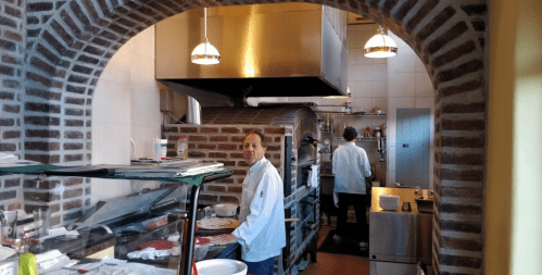 A view of a kitchen with two chefs working, featuring brick walls and stainless steel appliances.