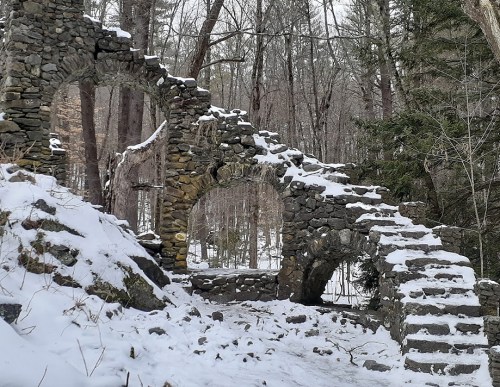 Snow-covered stone ruins with arches and stairs, surrounded by trees in a winter forest setting.