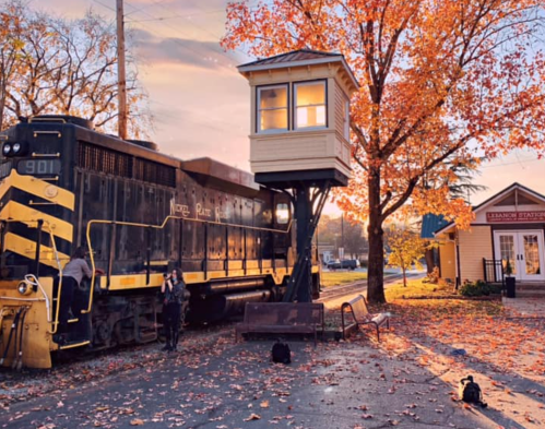 A vintage train near a control tower, surrounded by autumn leaves and a quaint station building in the background.