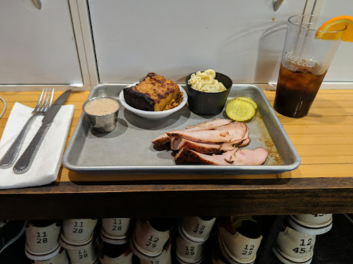 A metal tray with sliced meat, pickles, macaroni, cornbread, sauce, and a glass of cola with an orange slice.