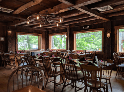 Cozy restaurant interior with wooden beams, empty tables, and large windows overlooking greenery.