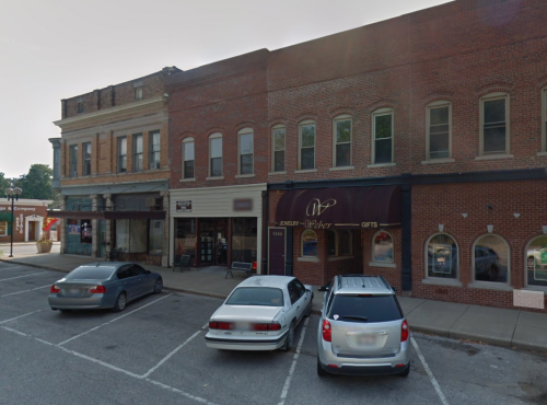 A street view of a small town with brick buildings, parked cars, and a jewelry store with an awning.