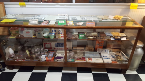 Display case filled with various vintage items, including bottles, papers, and containers, on a checkered floor.