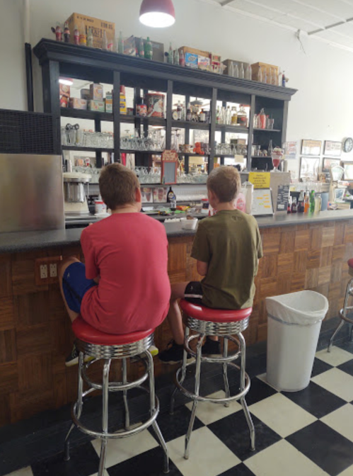 Two children sit on bar stools at a diner counter, facing a display of food and drinks. The floor has a black and white checkered pattern.