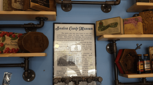 A wall display featuring shelves with vintage candy boxes and a sign about the Boston Candy Museum.