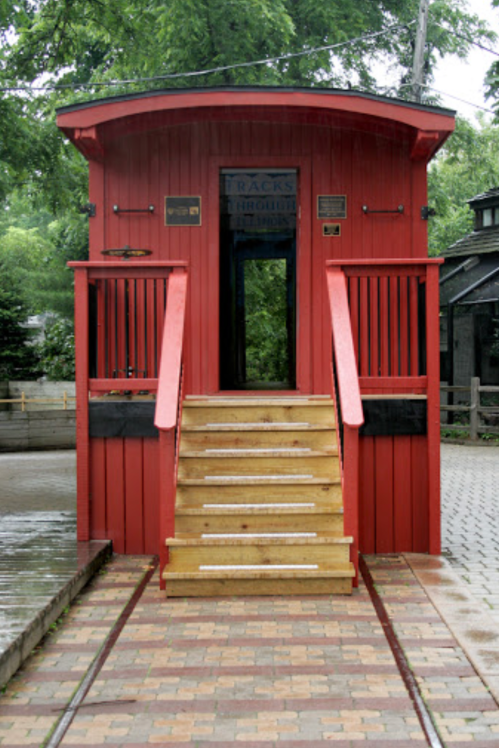 A red train caboose with stairs leading up, surrounded by greenery and brick pathways.