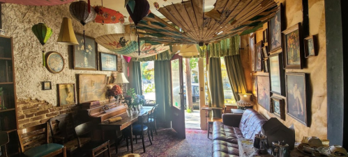 Cozy café interior with eclectic decor, including umbrellas, art, and a vintage piano, bathed in natural light.