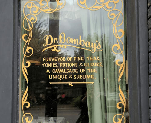 Sign on a window reading "Dr. Bombay's" with decorative text about fine teas, tonics, potions, and elixirs.