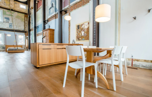 Modern kitchen with wooden cabinets, a dining table, and white chairs, featuring large windows and exposed brick walls.