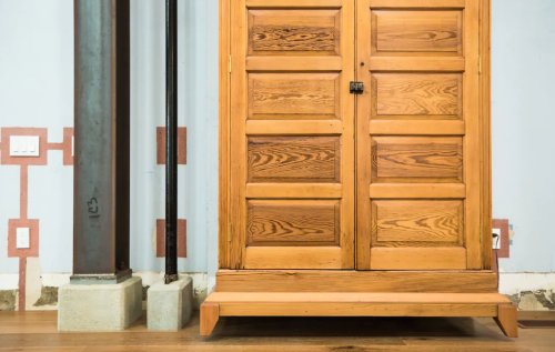 Wooden double doors with a simple handle, next to a metal support beam and concrete base, against a light blue wall.