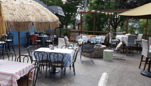 Outdoor dining area with tables, chairs, and a fire pit, surrounded by greenery and shaded by umbrellas.