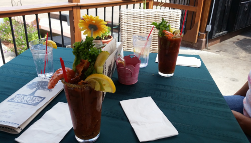 Three Bloody Mary cocktails with garnishes on a table, surrounded by water glasses and a sunflower centerpiece.
