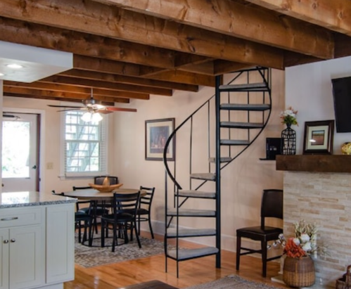 A cozy interior featuring a spiral staircase, wooden beams, a dining area, and a stone fireplace.