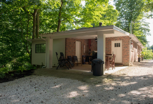 A cozy brick house with a covered porch, surrounded by lush green trees and a gravel pathway.