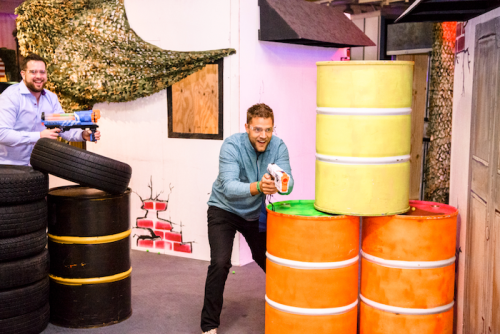 Two men play in a colorful indoor arena, hiding behind stacked barrels while holding toy blasters.