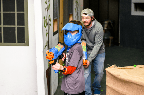 A child in a blue mask holds a toy blaster, while an adult crouches behind them in a play area.
