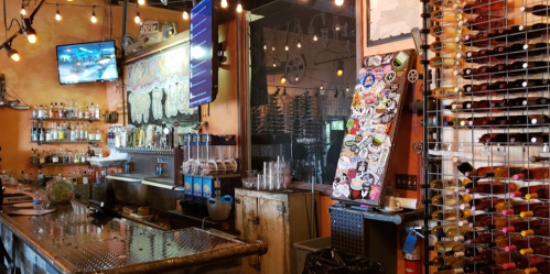 A cozy bar with a wooden counter, shelves of liquor, a TV, and a wall of wine bottles adorned with stickers.