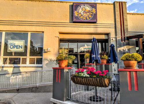 Exterior of a bakery with a sign, open umbrellas, and flower pots, showcasing a welcoming atmosphere.