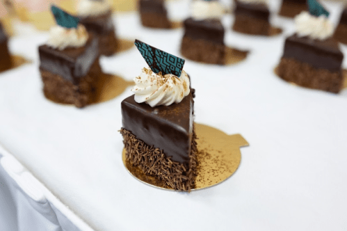 A close-up of a chocolate cake slice topped with cream and a decorative flag, placed on a gold base.
