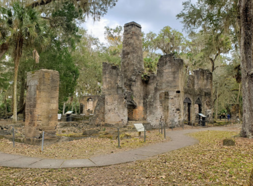 Ruins of an old stone structure surrounded by trees and grass, with informational signs nearby.