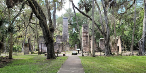 Ruins of stone structures surrounded by trees and moss, with a pathway leading through a lush green landscape.