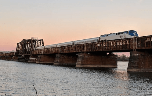 An Amtrak train crosses a rusty bridge over a river at sunset, with a clear sky in the background.