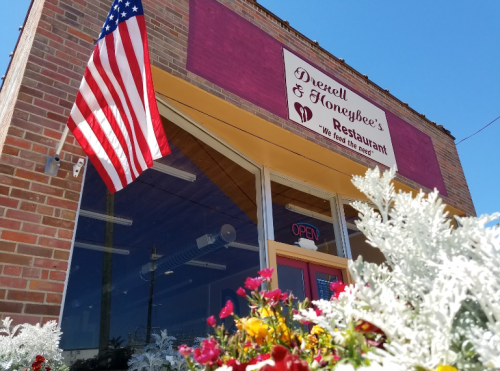 Exterior of Dwyell & Honeybee's Restaurant with an American flag and colorful flowers in front. Sign reads "Open."