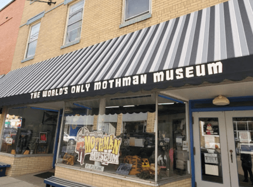 Exterior of the Mothman Museum, featuring a striped awning and a display of Mothman-themed merchandise in the window.