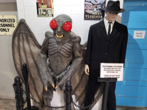 A mothman costume figure with red eyes and wings stands next to a mannequin in a suit, displayed behind a rope barrier.