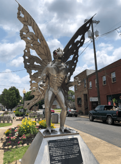 A metallic statue of a humanoid figure with butterfly wings, standing in a town square surrounded by flowers.