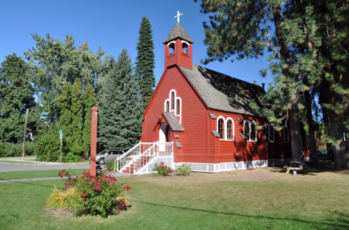 A red church with a steeple, surrounded by trees and a grassy area, under a clear blue sky.
