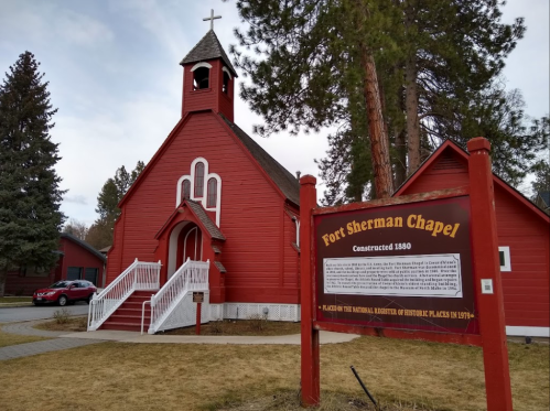 Red wooden chapel with a bell tower, sign reading "Fort Sherman Chapel, Constructed 1880," surrounded by trees.