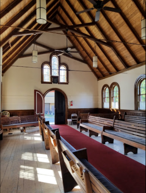 Interior of a church with wooden beams, benches, and a red carpet leading to an arched doorway. Natural light filters in.