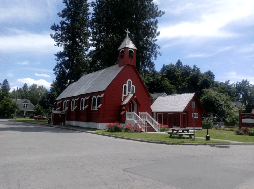 A red church with a steeple, surrounded by trees and flowers, on a sunny day with blue skies.