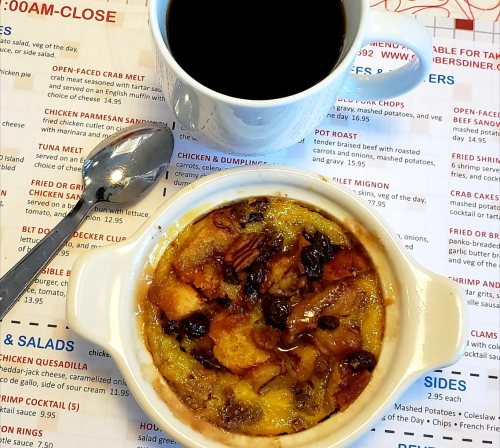 A bowl of bread pudding with raisins, a spoon, and a cup of coffee on a menu background.