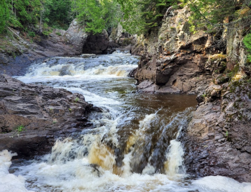 A serene river flows over rocky terrain, surrounded by lush green trees and cascading waterfalls.