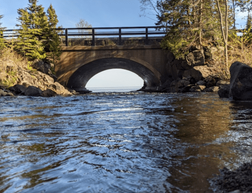 A serene river flows under a stone bridge, surrounded by trees and rocky banks, with a clear blue sky above.