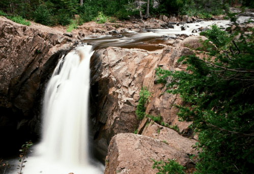 A serene waterfall cascades over rocky terrain, surrounded by lush greenery and flowing water.