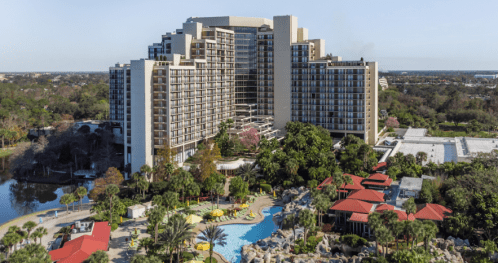 A large hotel building with multiple floors overlooks a pool area surrounded by palm trees and colorful cabanas.