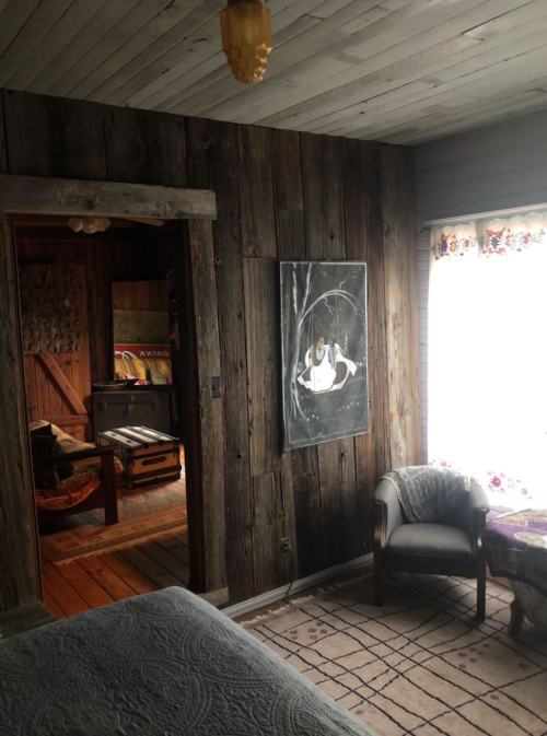 Cozy room with wooden walls, a painting, and a window with floral curtains, featuring a chair and a glimpse of another room.