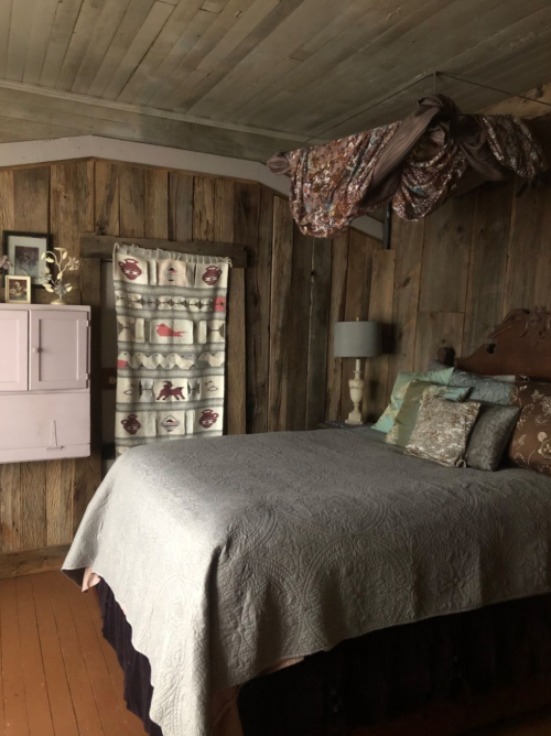 Cozy bedroom with a wooden wall, a bed with decorative pillows, and a vintage quilt, featuring soft lighting and decor.