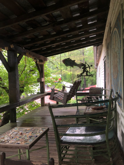 A rustic porch with a swing, wooden chairs, and a table, surrounded by greenery and a decorative wall.