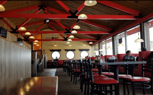 Interior of a restaurant featuring red beams, round windows, and black chairs with red cushions. A few patrons are seated.
