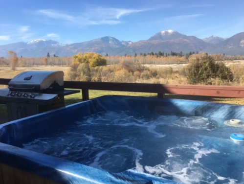 A hot tub on a deck with a grill, overlooking a scenic view of mountains and a clear blue sky.