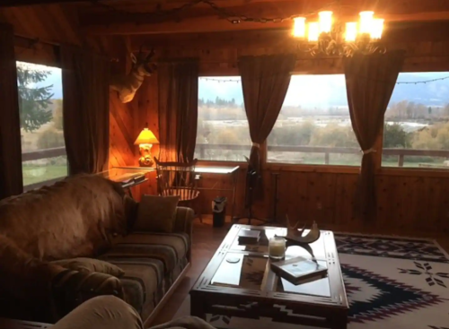 Cozy cabin interior with a sofa, coffee table, and large windows showcasing a scenic view. Warm lighting adds ambiance.